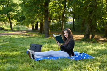 beautiful long-haired woman reading a green book in nature sitting on the grass with a blanket and her bag with trees in the background. teenager looking at a literary work in the park smiling