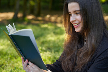 beautiful long-haired woman reading a green book in nature sitting on the grass with a blanket and her bag with trees in the background. teenager looking at a literary work in the park smiling