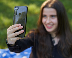 young woman with long hair in a black shirt and white pants sitting on a blanket of stars on the grass in a park with her bag using her phone to take pictures and video conferencing. influencer nature