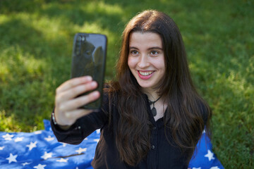 young woman with long hair in a black shirt and white pants sitting on a blanket of stars on the grass in a park with her bag using her phone to take pictures and video conferencing. influencer nature