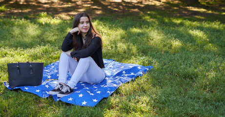 Pretty young woman with long hair in a black shirt and white pants sitting on a blanket of stars on the grass in a park with her bag looking to the sides. teenager resting in nature with blue blanket