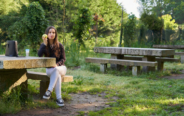 beautiful young woman in a black shirt sitting on a stone table eating an apple in nature with her mobile, a coffee and her bag with trees in the background. vegan teen taking a break in the park