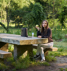 beautiful young woman in a black shirt sitting on a stone table eating an apple in nature with her mobile, a coffee and her bag with trees in the background. vegan teen taking a break in the park