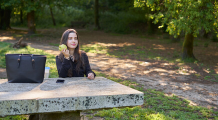 beautiful young woman in a black shirt sitting on a stone table eating an apple in nature with her mobile, a coffee and her bag with trees in the background. vegan teen taking a break in the park