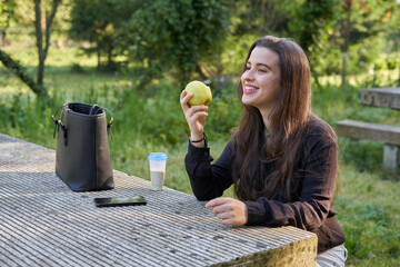 beautiful young woman in a black shirt sitting on a stone table eating an apple in nature with her mobile, a coffee and her bag with trees in the background. vegan teen taking a break in the park