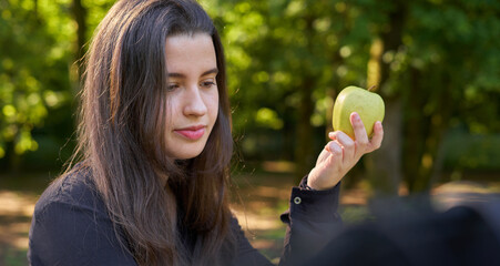 pretty young woman in black shirt sitting at a stone table looking at her phone and eating an apple while having coffee with trees in the background. vegan teen taking a break with her cell phone