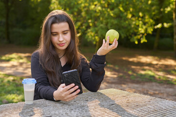 pretty young woman in black shirt sitting at a stone table looking at her phone and eating an apple while having coffee with trees in the background. vegan teen taking a break with her cell phone