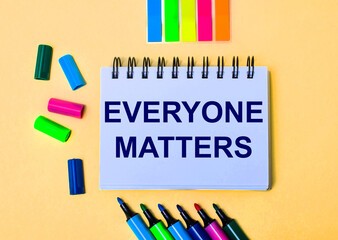 The words EVERYONE MATTERS written in a white notebook on a beige background near multi-colored markers
