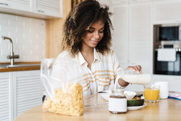 The smiling black woman pouring a milk into a breakfast plate while sitting at the table in the kitchen