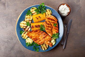 Grilled chicken breast with corn and zucchini on a blue plate top view. Summer dish with grilled chicken and vegetables.