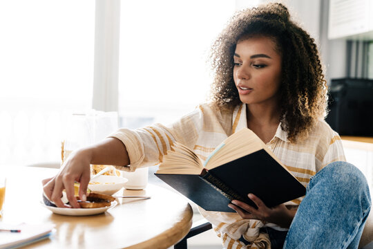 The Side View Of A Woman Holding A Book In Her Hands And Reaching For A Cookie While Sitting In The Kitchen