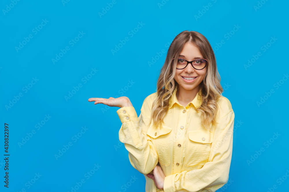 Wall mural A young caucasian beautiful happy smiling blonde student promoter woman in a yellow shirt and glasses holds her hand demonstrating empty copy space ads promo offer on a bright color blue background