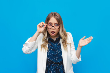 A young caucasian shocked surprised pretty blonde business woman in a white jacket and glasses with open mouth holding glasses in her hand on bright color blue background. Shocking news, starled face