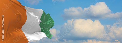Large flag of Cote d'lvoire  waving in the wind on flagpole against the sky with clouds on sunny day. 3d illustration