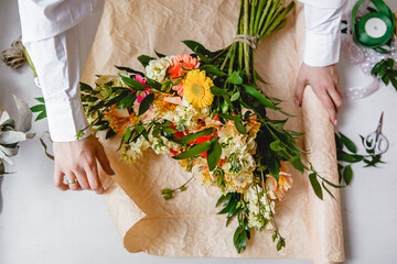 a female florist in a white shirt wraps a bouquet of fresh flowers in paper. Top view - obrazy, fototapety, plakaty
