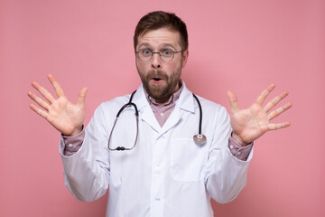 Joyful doctor with a stethoscope around neck spread his hands in surprise and looks at the camera with mouth open. Pink background.