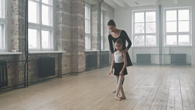 Full shot of gracious little girl performing ballet movements to female trainer during lesson at choreography class