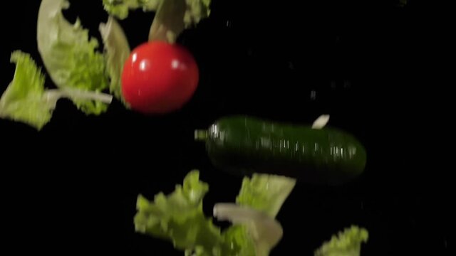 Close up on the fresh tomatoes, cucumbers and lettuce tossed up and spinning with water splashes on a black background in slow motion shot, water drops on vegetables