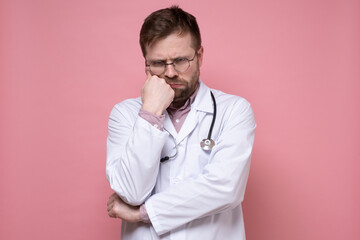 Frowning, a doctor in round glasses, a white coat and with a stethoscope around neck looks sternly and displeased with fist to face. 