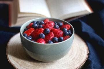 Bowl of blueberries and strawberries on wooden tray and open book. Selective focus, dark textile background.