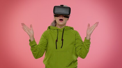 Senior granny gray-haired woman using virtual reality futuristic technology VR app headset helmet to play simulation 3D 360 video game, drawing. Pink studio background. Elderly grandmother pensioner