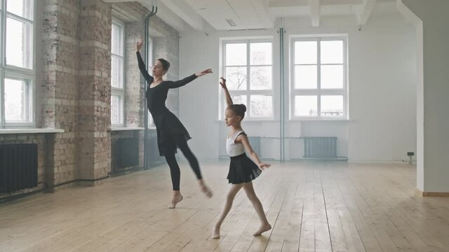 Full shot of young professional ballerina in black ballet dress dancing together with pretty little girl, moving synchronically at dance class