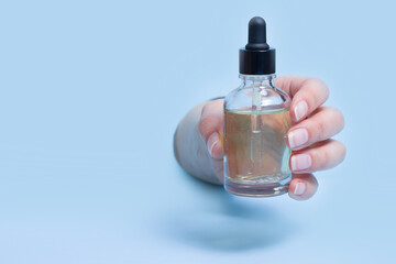 Female hand holds a glass dropper bottle on blue background. Medical or cosmetic oil