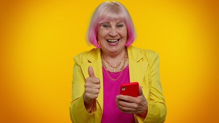 Happy excited joyful elderly granny gray-haired woman use mobile cell phone typing browsing say wow yes, found out great big win news doing winner gesture. Senior old grandmother on orange background