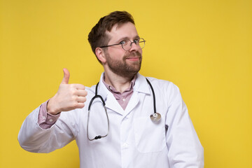 Cute, contented doctor shows an approving gesture, thumbs up and smiles friendly while looking into the camera. 