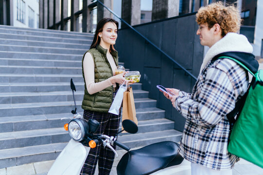 Red Haired Curly Caucasian Man Courier On Scooter Delivering Food In Holding Food Bag In Front Steps Of Modern Building And Brunette Smiling Woman Accepting A Delivery Of Bag From Deliveryman.