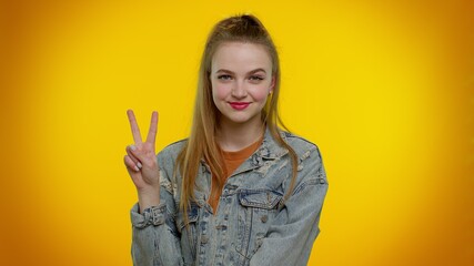 Pretty stylish teen girl in denim jacket showing victory sign, hoping for success and win, doing peace gesture, smiling with kind optimistic expression. Young adult woman on yellow wall background