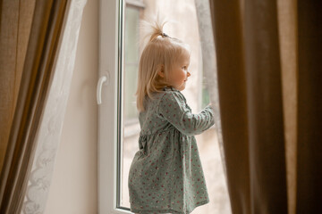 Cute baby blonde girl in vintage dress stands by the window of the house