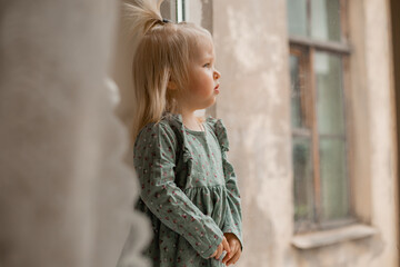 Fototapeta na wymiar Cute baby blonde girl in vintage dress stands by the window of the house
