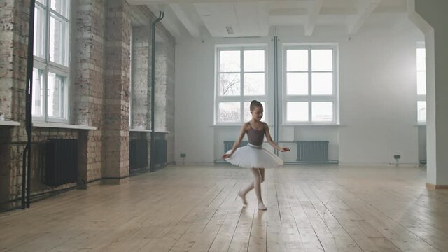 Full shot of cute little ballerina in white tutu dancing alone in spacious loft-style dance class