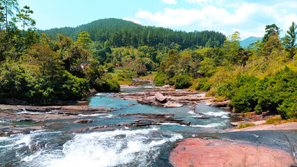 Obraz premium Unique shot of clean water cascading nicely over a cool stone plain near a scenic greenish pine forest on a sunny day - One of the major tourist attracted places in Sri Lanka.