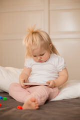 Fototapeta premium Baby blonde girl plays with the phone at home on the bed. Children and technology