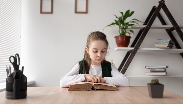 School girl sitting indoors and reading a thick old book quickly turning the pages. A gentle pensive child learns online remotely at home, completing the teacher's assignments.