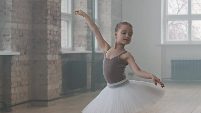 Medium portrait of cute 9-year-old ballerina posing to camera standing in ballet pose at dance class wearing white tutu