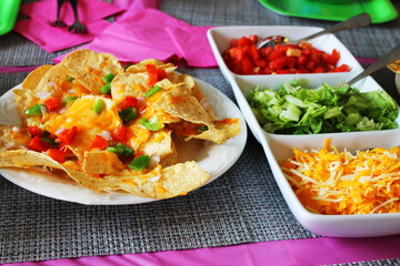 Close-up of a table set for a party, with nachos, a dish of taco toppings, a pink table cloth and napkins, place mats and paper plates.