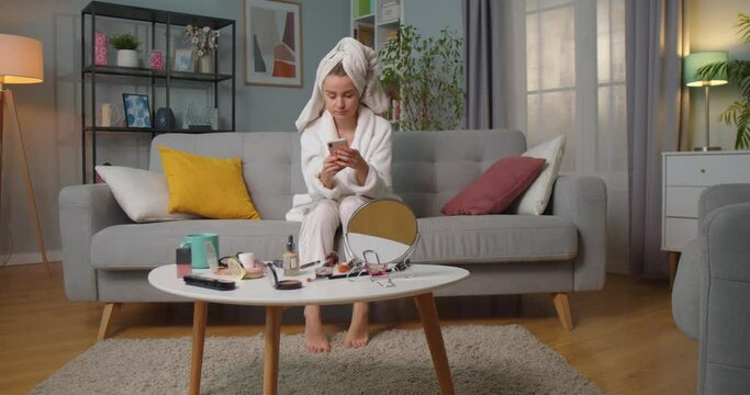 Middle plan of woman in bathrobe and towel sitting on sofa in living room texting, chatting, reading messages, smiling using smartphone and put her legs on couch. Makeup kits on table in living room.
