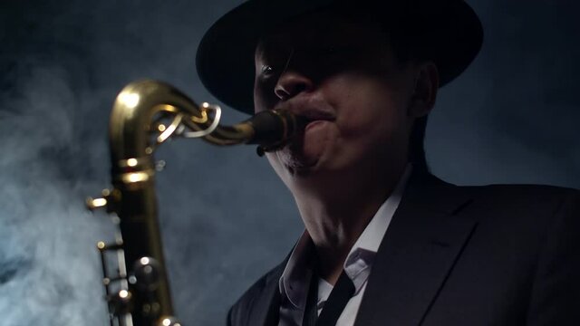 Man saxophonist in a hat and suit plays the saxophone in a smoky studio. Adult male musician blows close-up saxophone. Concept musical education.