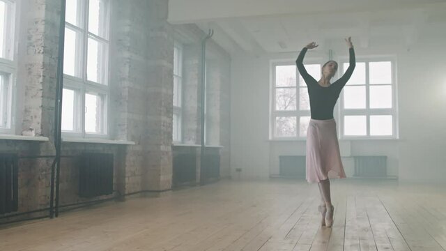 Full shot of talented young ballerina dancing gracefully in empty smoke-filled dance studio enjoying process
