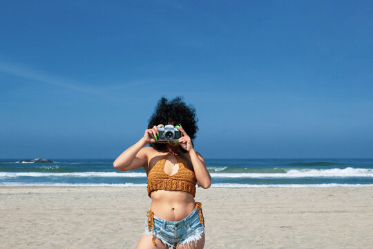 beautiful and sensual Mexican woman with curly hair taking pictures on the beach with old camera
