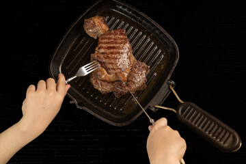 The child holds a cutlery fork and a knife and cuts them steak in a hot frying pan. Black background