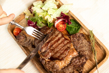 Ready grilled steak on a wooden plate, a portion of fresh salad. Children's hands cut meat.