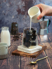 Intense black coffee ice cube in a glass with hand pouring milk from pot to make cold brew latte with coffee grains in background, easy homemade beverage to refreshing during hot summer 