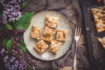 Pieces of rhubarb crumble cake on a plate on a wooden background, decorated with lilac flowers, top...