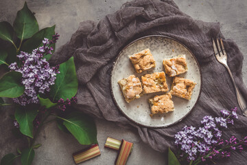 Pieces of rhubarb crumble cake on a plate on a gray background, decorated with lilac flowers, top view
