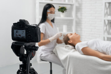 Naklejka premium Side view of brunette beautician in protective mask and special white uniform doing special procedure for hair for young woman in camera. Concept of recording on camera in beauty salon. 