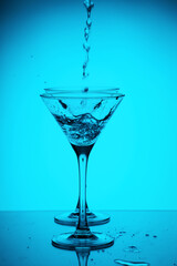 Studio photo of two triangular shape glass with pouring pure water into them. Transparent martini glass on light blue background. Vertical image with alcohol